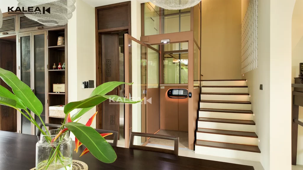 Pintu lift rumah Kalea terbuka dengan shaft kaca di sebelah tangga