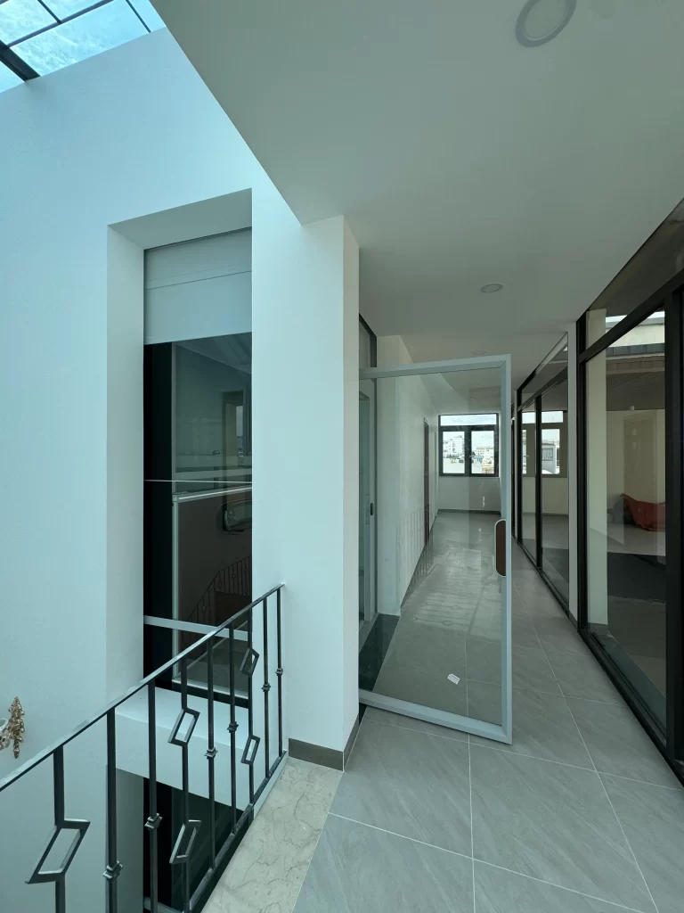Detail lift rumah Kalea dalam shaft beton dengan pintu kaca di koridor lantai atas