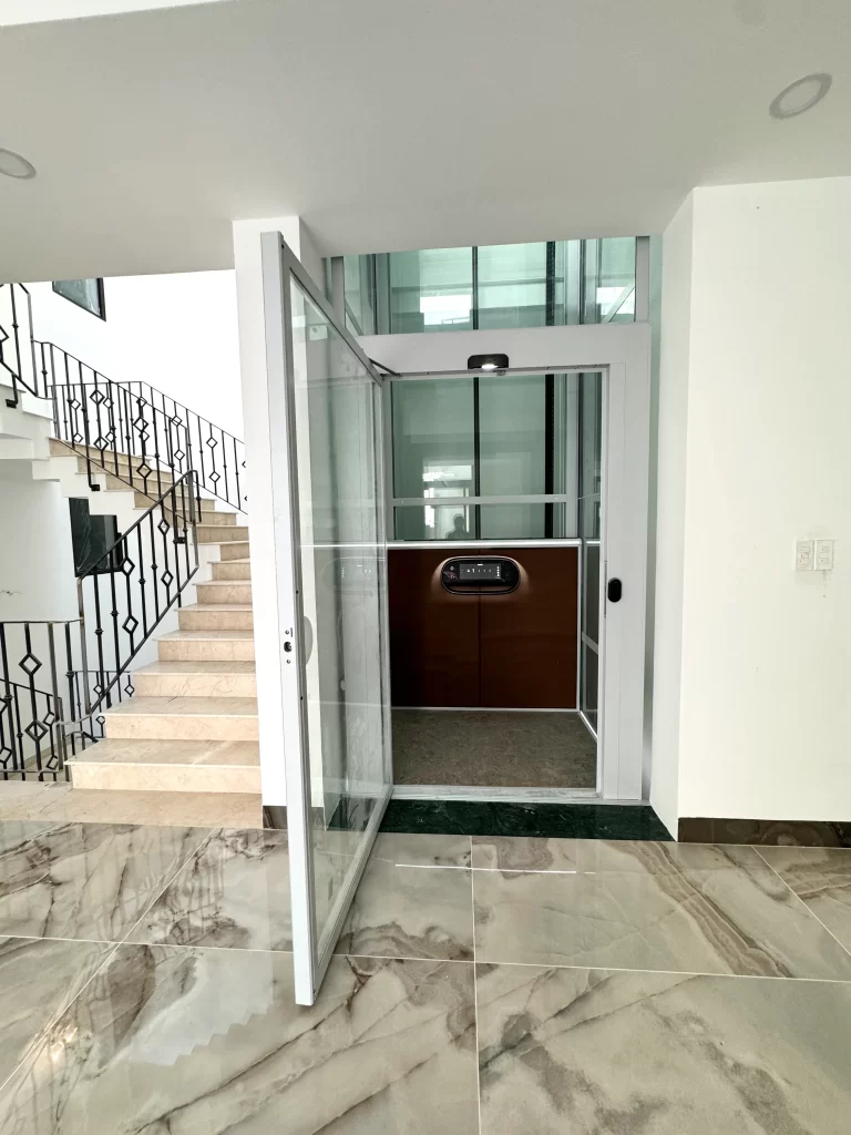 Lift rumah Kalea dengan pintu kaca terbuka dan shaft putih di samping tangga