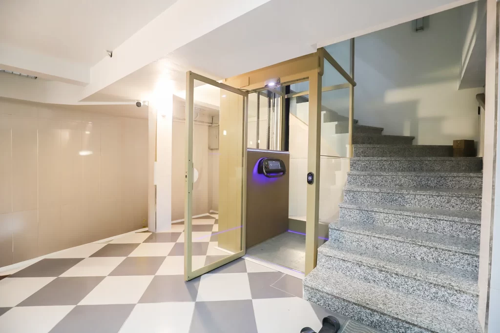 Lift rumah Kalea dengan pintu kaca terbuka, terpasang di samping tangga dalam hunian bertingkat