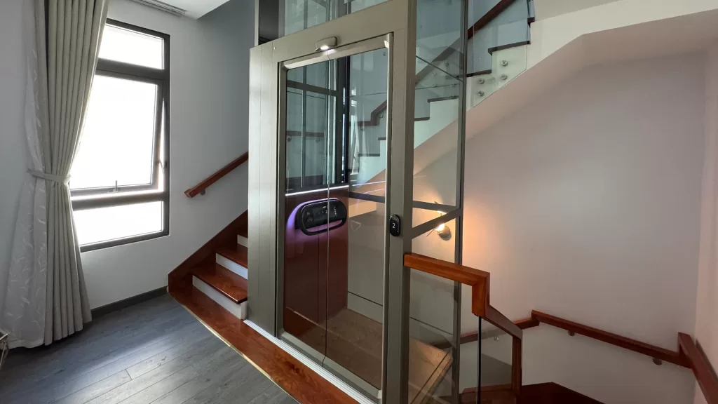Lift rumah Kalea X50 Limited di area tangga rumah modern, menunjukkan bagaimana lift rumah mulai terasa penting untuk hunian bertingkat yang mengutamakan kenyamanan akses harian
